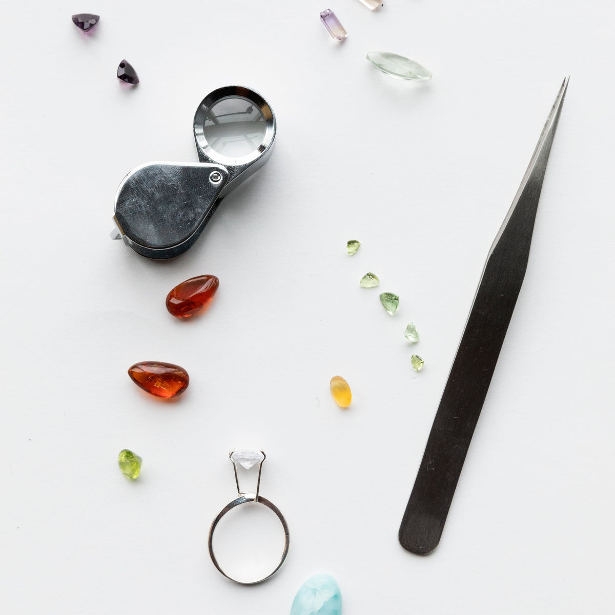 Jewelry tools including a magnifying glass, tweezers, and various gemstones on a white background